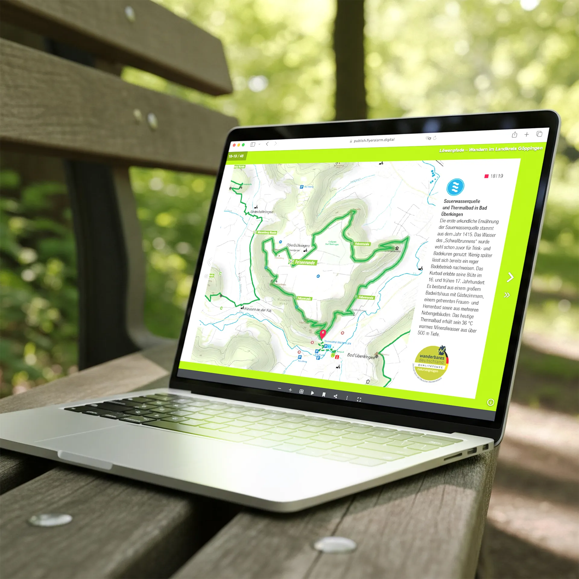 Laptop auf einer Holzbank im Freien mit geöffnetem Fenster, das eine digitale Wanderkarte des Löwenpfad-Projekts im Landkreis Göppingen zeigt. Die Karte markiert Wanderrouten und Points of Interest wie die Sauerwasserquelle und das Thermalbad in Bad Überkingen. Im Hintergrund grüne Blätter und Lichtreflexe.